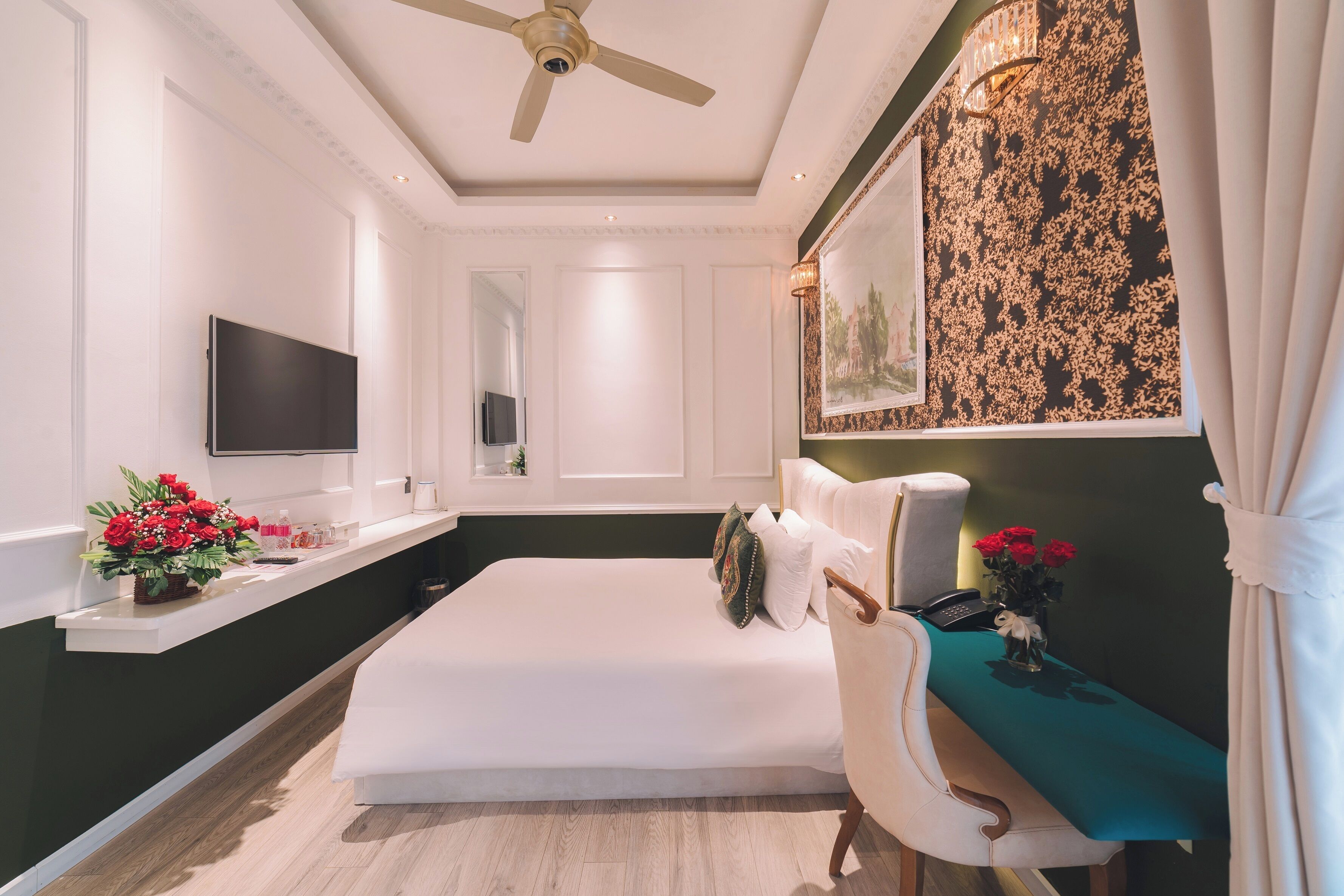 A hotel room with a bed, a desk with a chair, a flat-screen TV, a ceiling fan, and a painting on the wall.