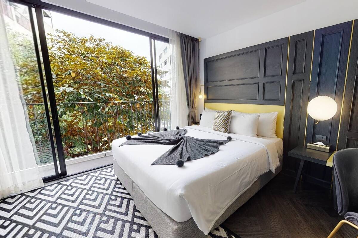 A hotel room with a large bed, a desk with a lamp, and a view of trees through the window.