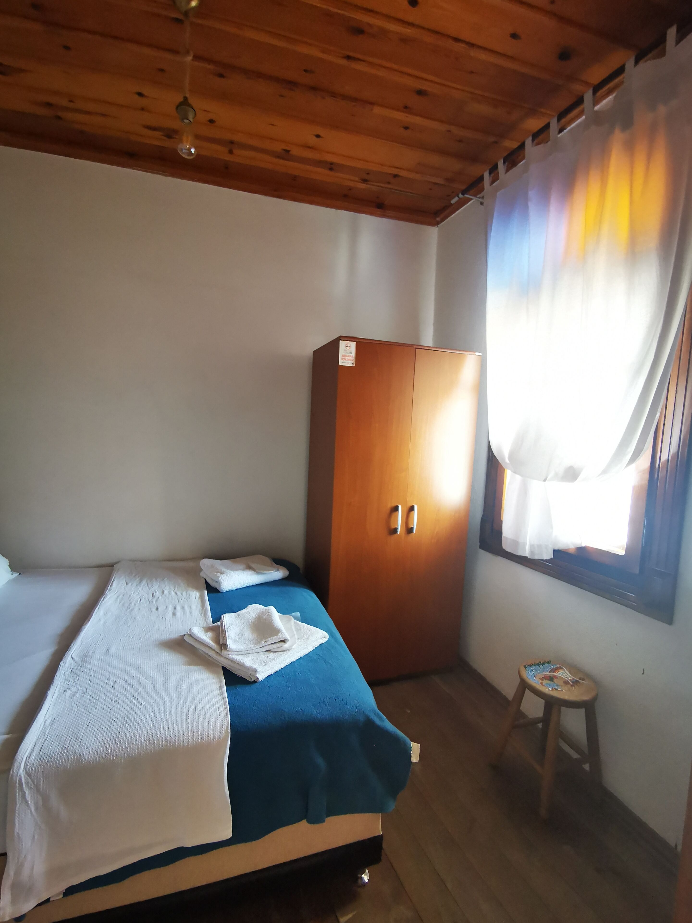 Ein Zimmer mit einem Bett, einem Kleiderschrank, einem kleinen Hocker und einem Fenster mit Vorhängen.
