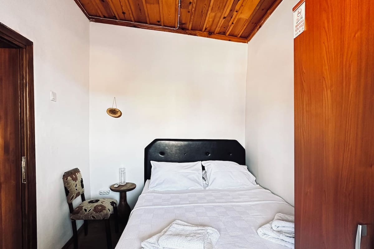 A bedroom with a wooden ceiling, a bed with white bedding, a small round table with a chair, and a door on the right.
