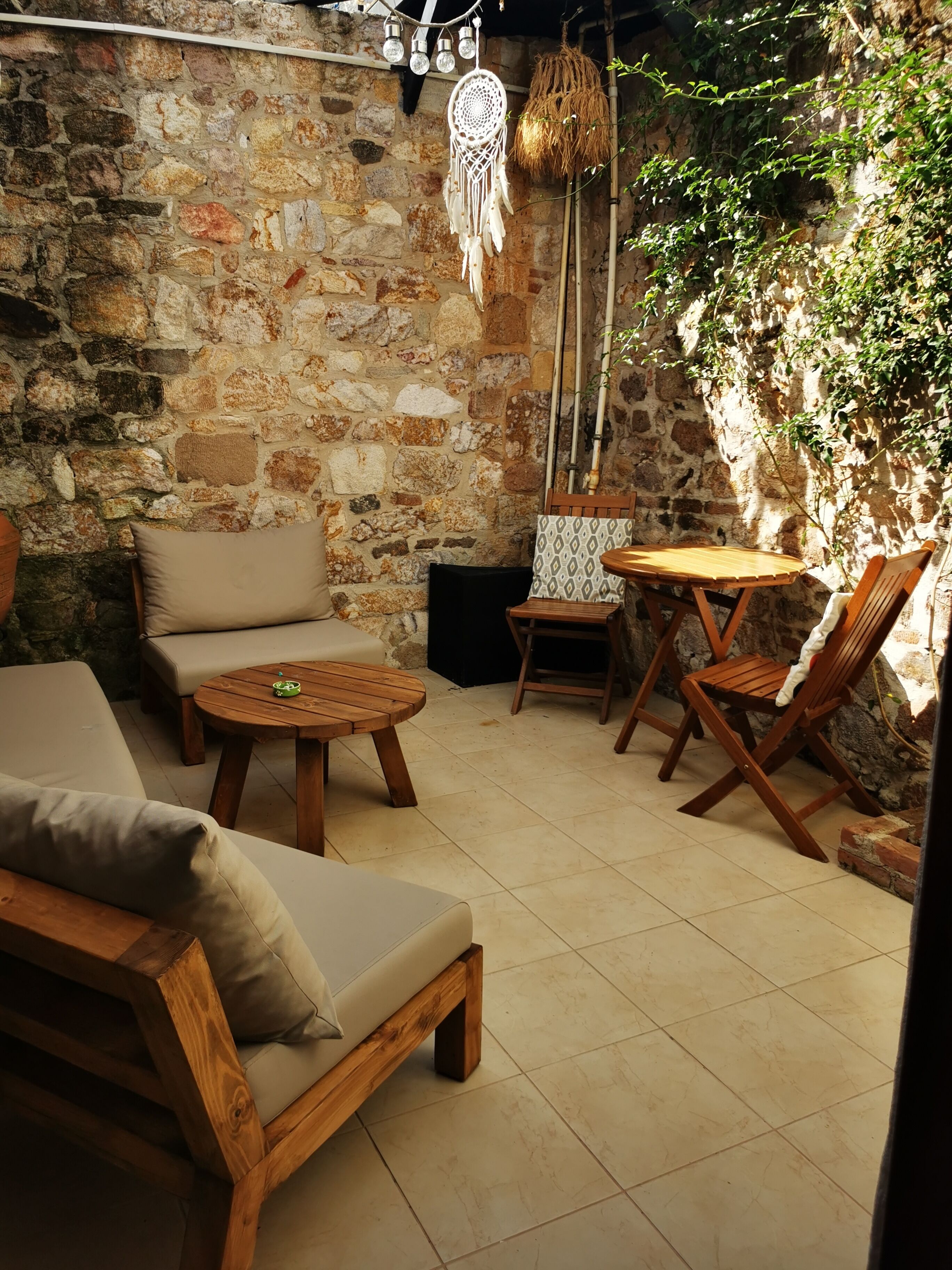 A cozy outdoor seating area with a stone wall, wooden furniture, and a dreamcatcher hanging above.