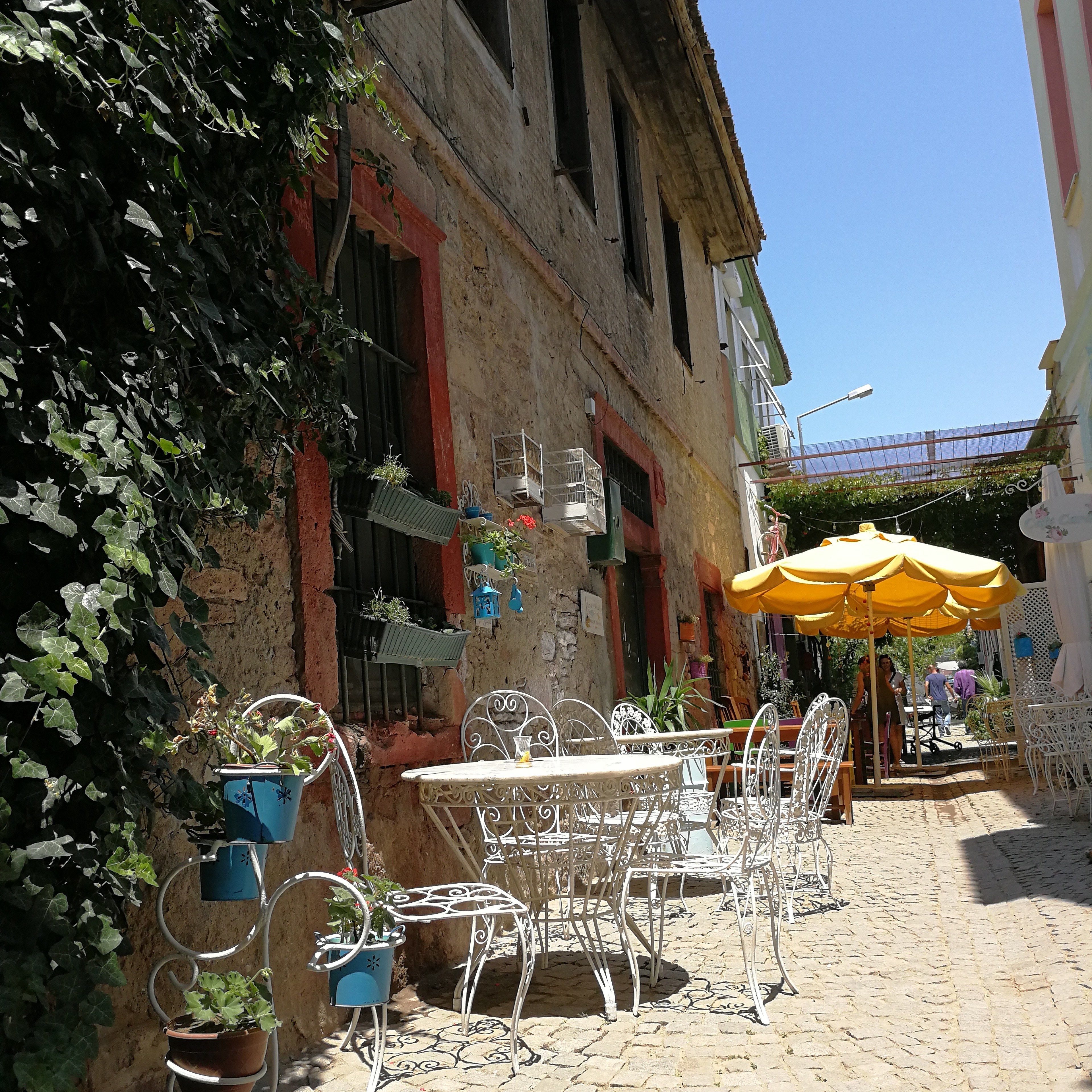 An outdoor cafe with white metal chairs and tables, surrounded by potted plants and a yellow umbrella.