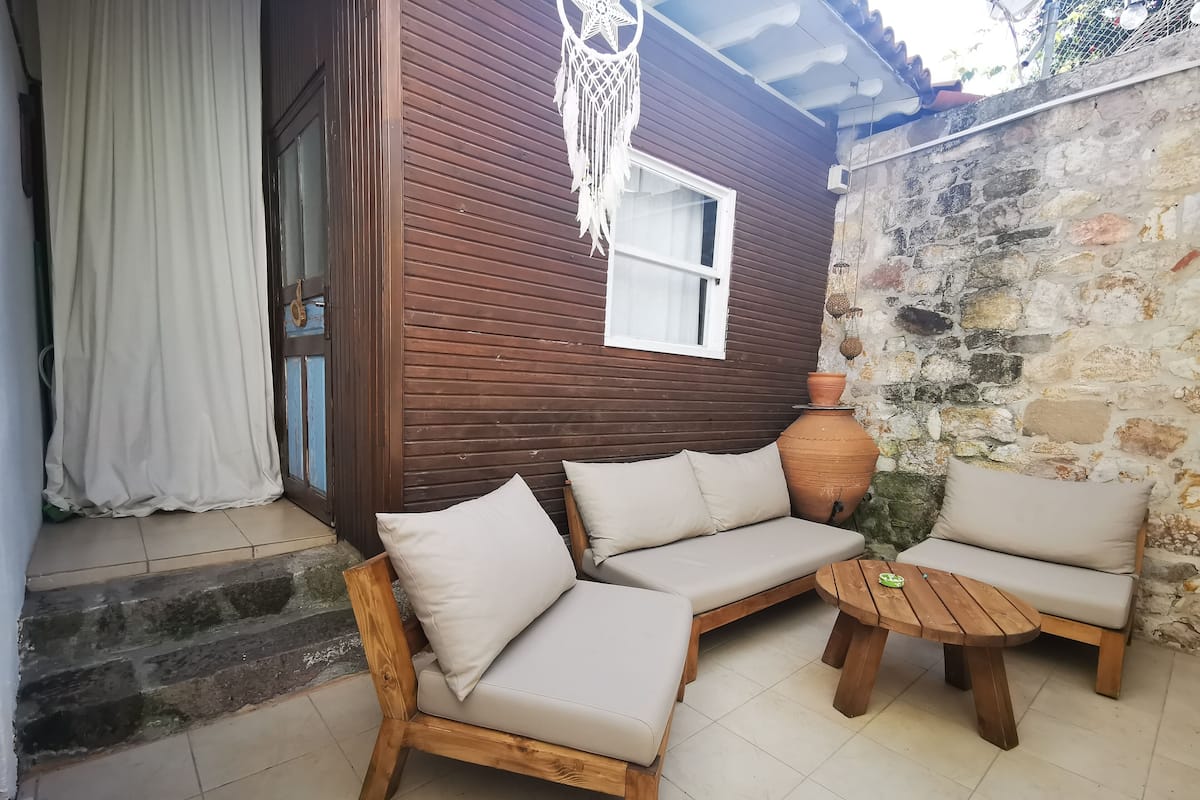 A wooden house with a brown exterior, a white door, and a white curtain. Inside, there is a beige sofa with cushions, a round wooden table, and a large terracotta pot.