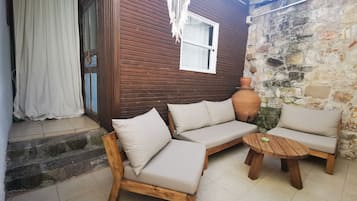 Une maison en bois à l’extérieur brun, avec une porte blanche et un rideau blanc. À l’intérieur, on trouve un canapé beige avec des coussins, une table ronde en bois et un grand pot en terre cuite.