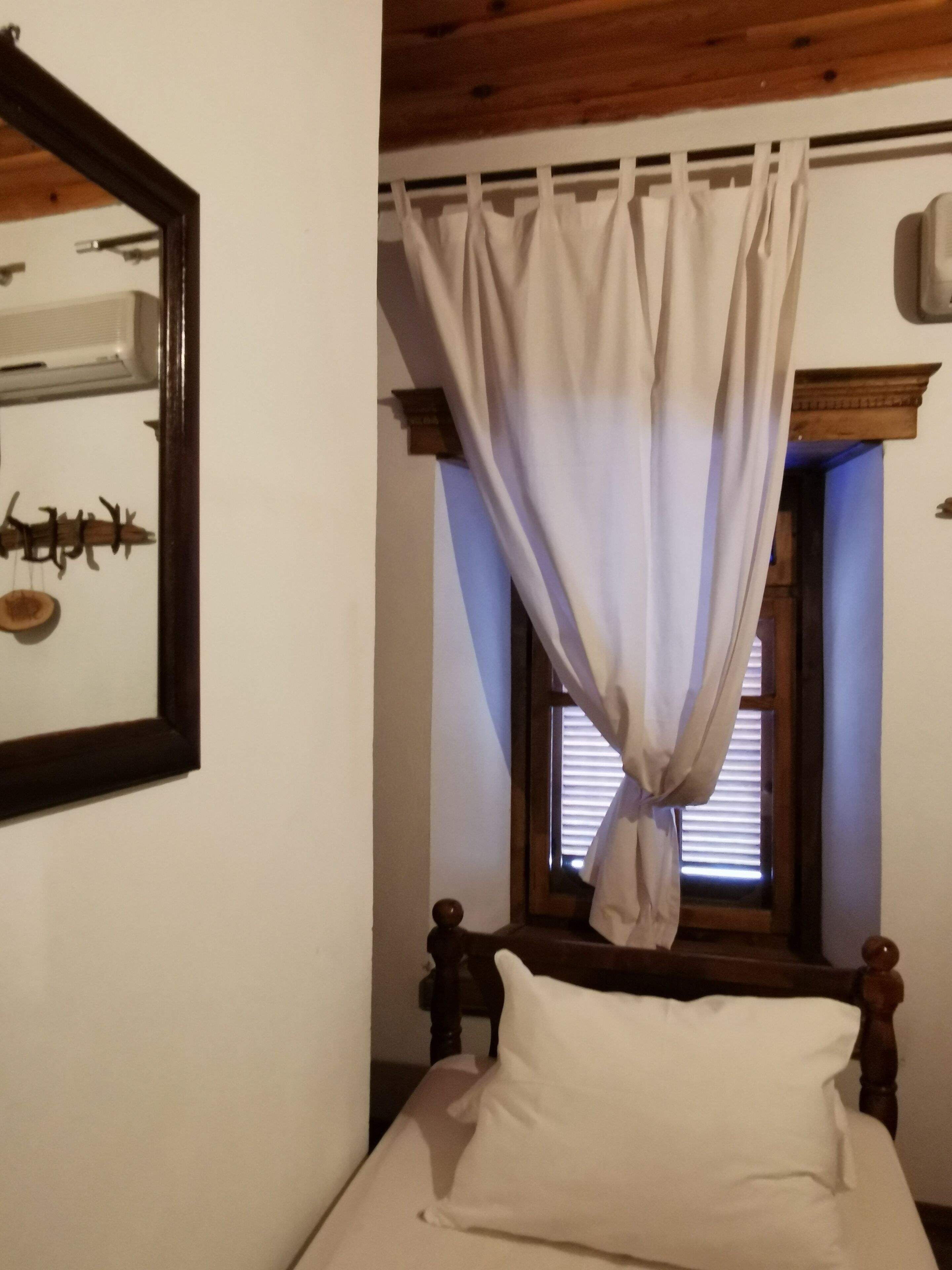 A bedroom with a bed, wooden headboard, a window with curtains, and a wall-mounted mirror.