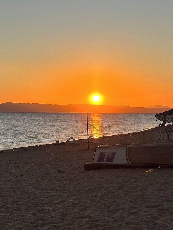 A beach scene with a sunset, a beach umbrella, and a chair. - Sahmeran Pansiyon (Ayvalik)