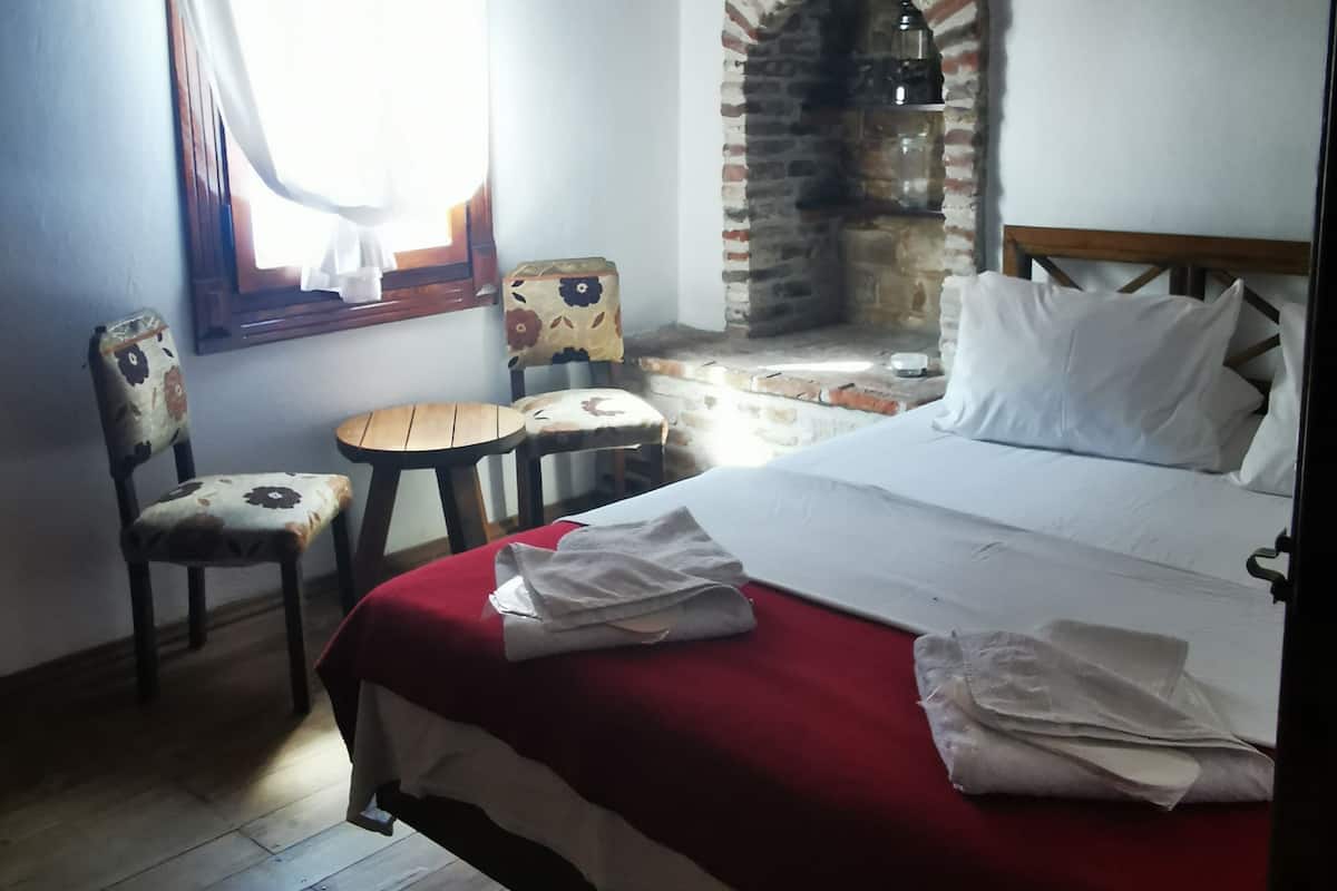 A bedroom with a brick fireplace, a bed with white linens, a wooden ceiling, and a small table with chairs.
