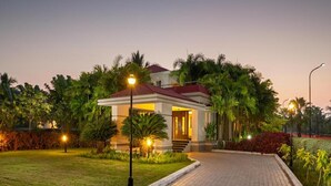 A well-lit, modern building with a red roof, surrounded by lush greenery and a paved pathway leading to the entrance. - Sunray Village Resort (Bhogopuram)