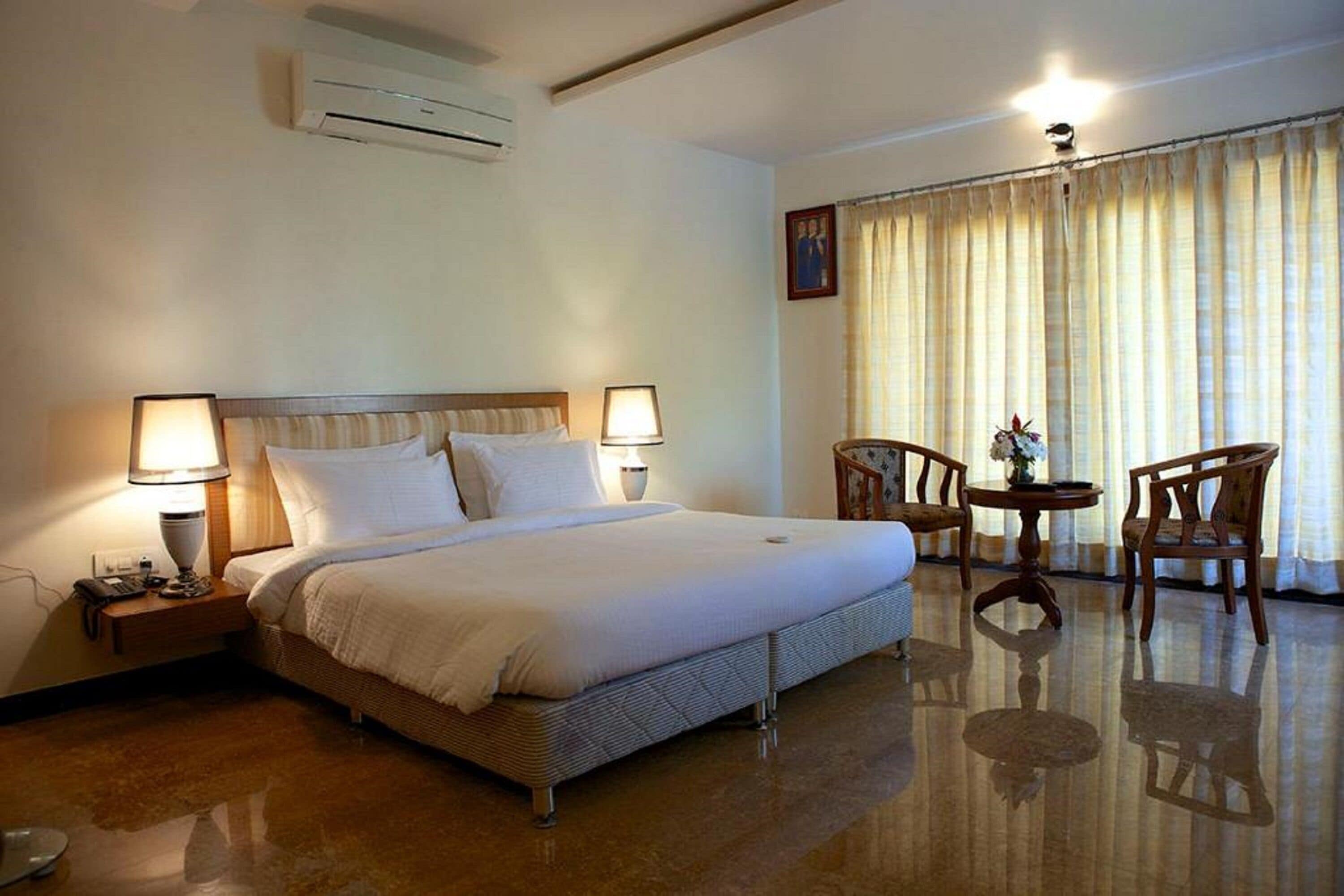 A hotel room with a large bed, two bedside lamps, a small round table with chairs, and a window with curtains.