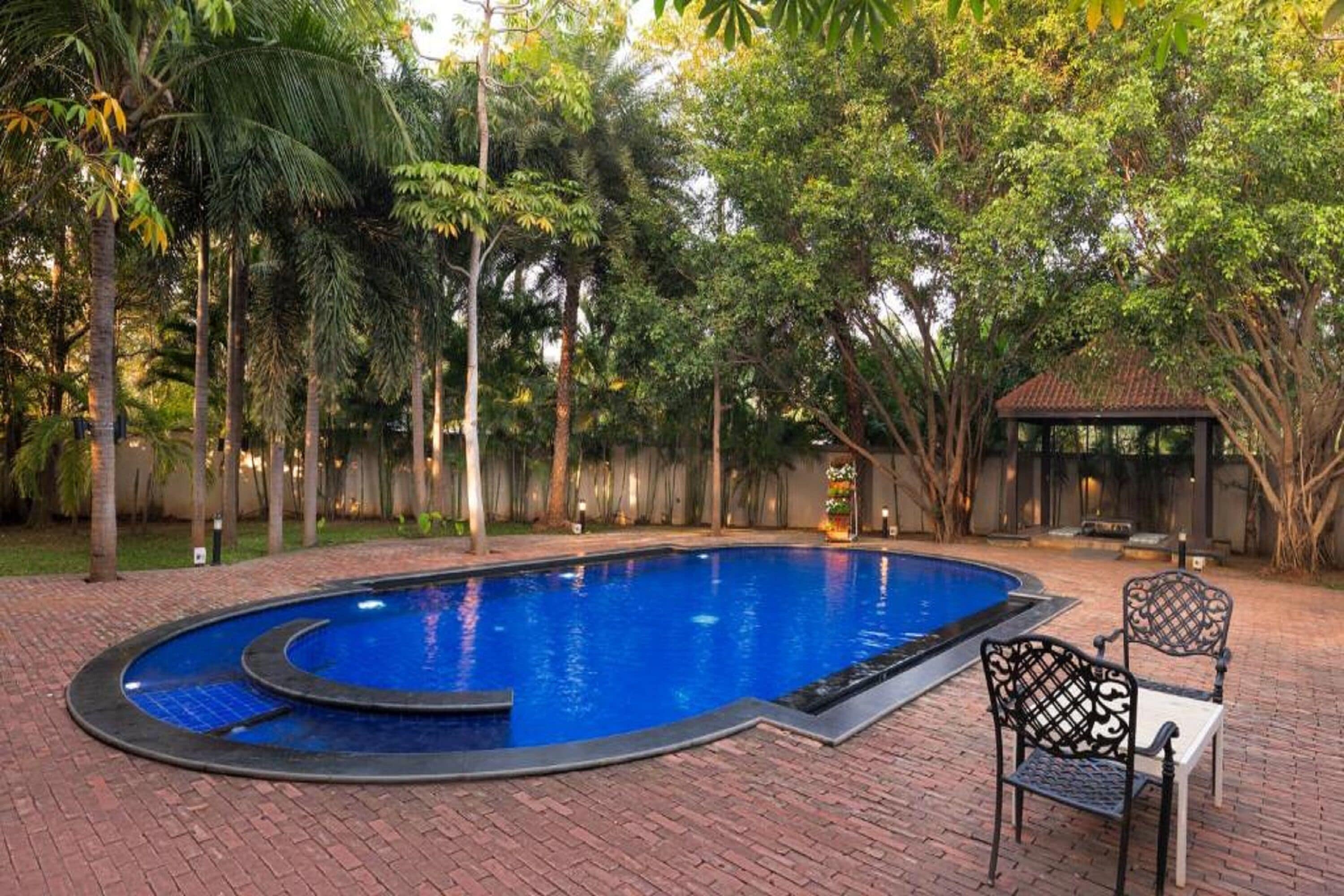 A swimming pool with a curved edge, surrounded by a brick patio. There is a gazebo and a chair in the background.