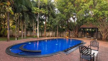 A swimming pool with a curved edge, surrounded by a brick patio. There is a gazebo and a chair in the background.