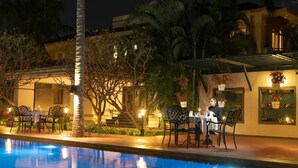 A poolside dining area with tables and chairs, a pool, and a building with a balcony in the background. - Sunray Village Resort (Bhogopuram)