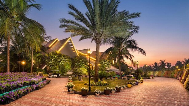 A landscaped garden area with palm trees, a paved walkway, and a building with a distinctive roof design. - Sunray Village Resort (Bhogopuram)