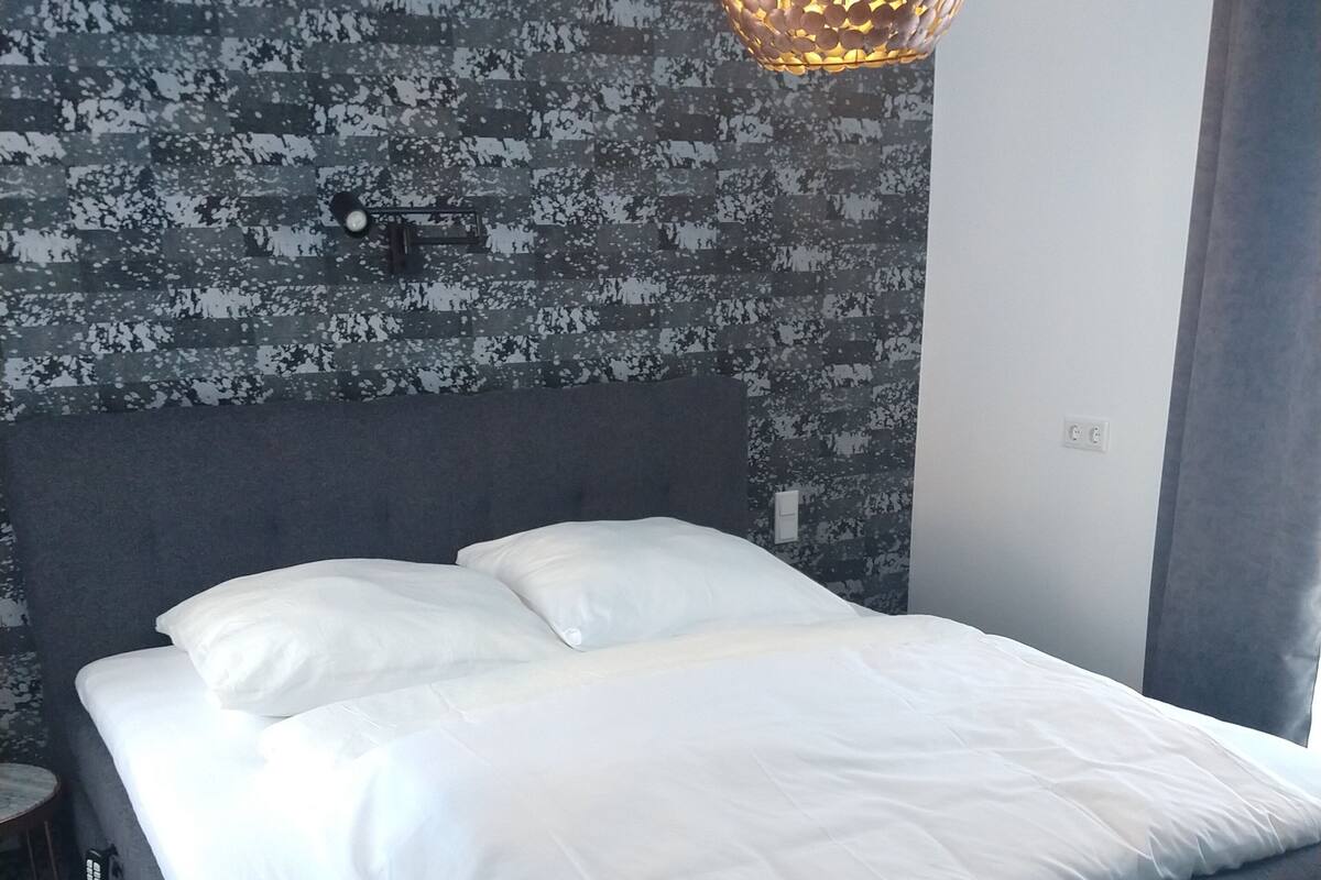 A neatly made bed with white linens in a room with a textured wall and a hanging light fixture.