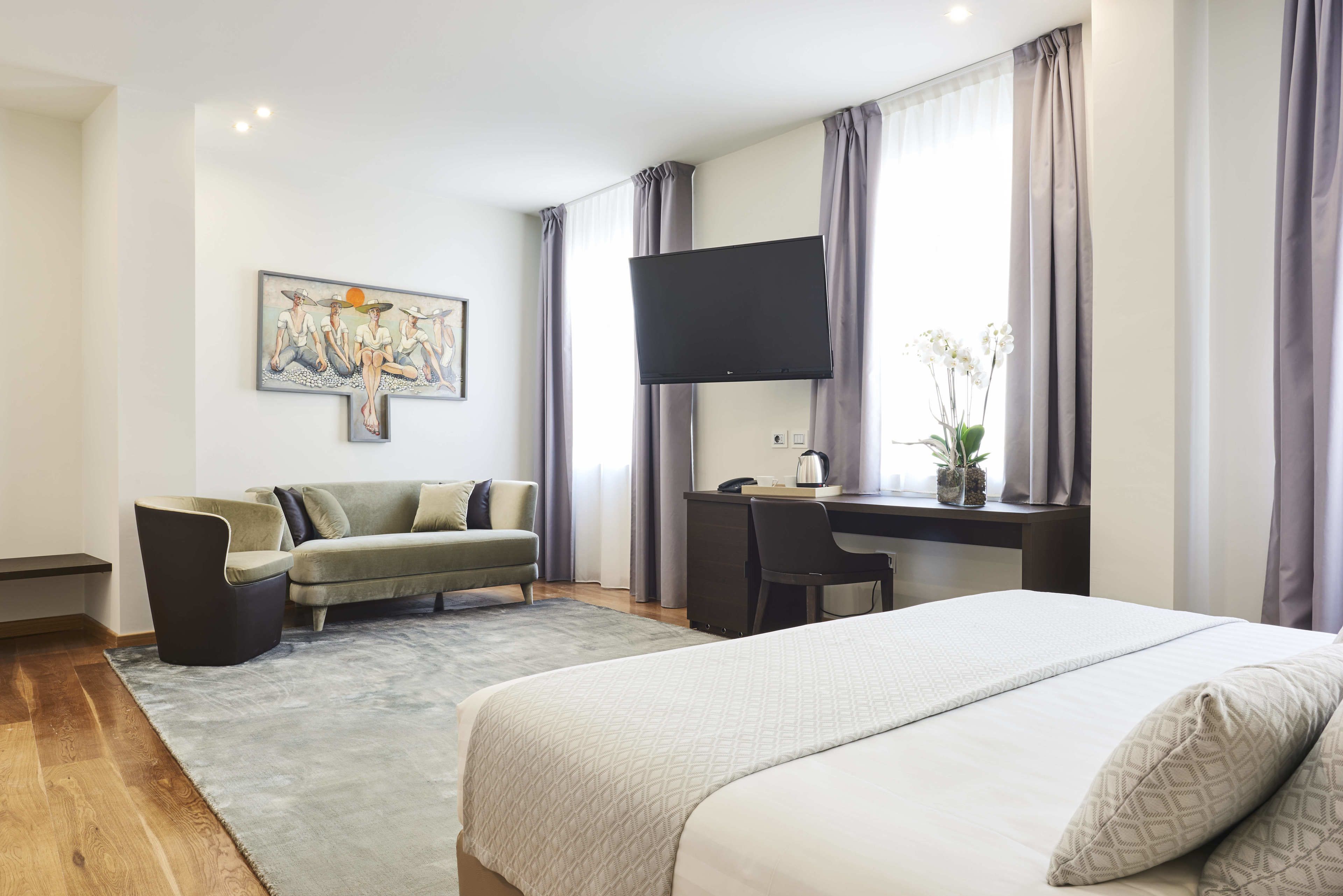 A modern hotel room with a bed, a desk with a computer, a sofa, and a flat-screen TV.