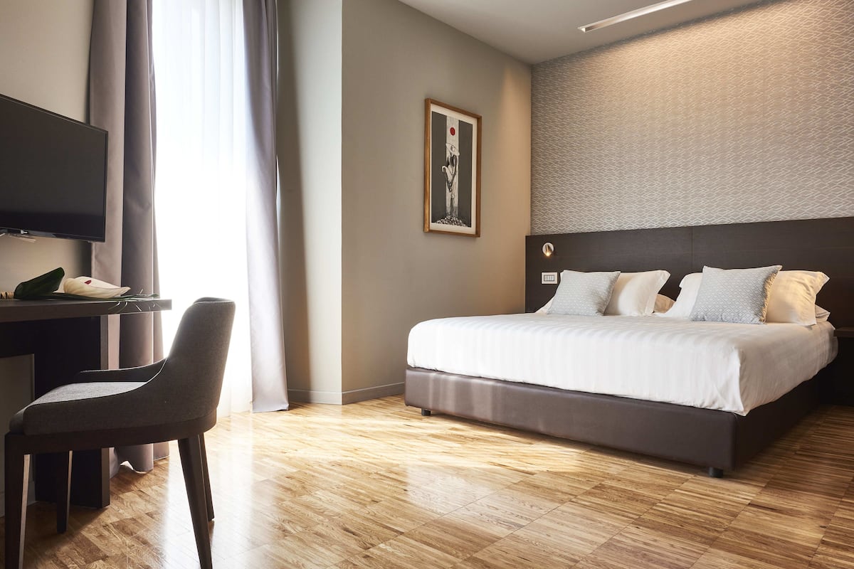 A modern hotel room with a bed, a desk with a chair, a television, and a framed picture on the wall.