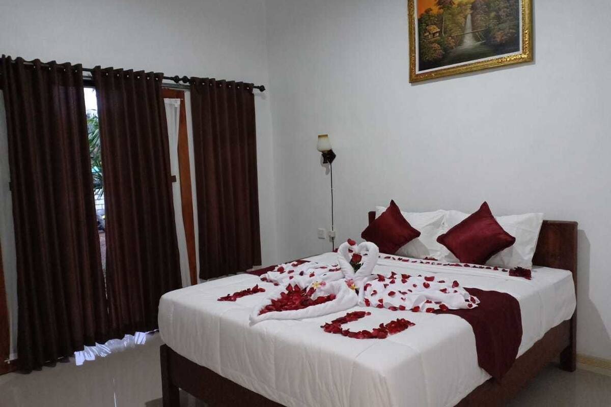 A hotel room with a bed, red curtains, a painting on the wall, and a nightstand with a lamp.