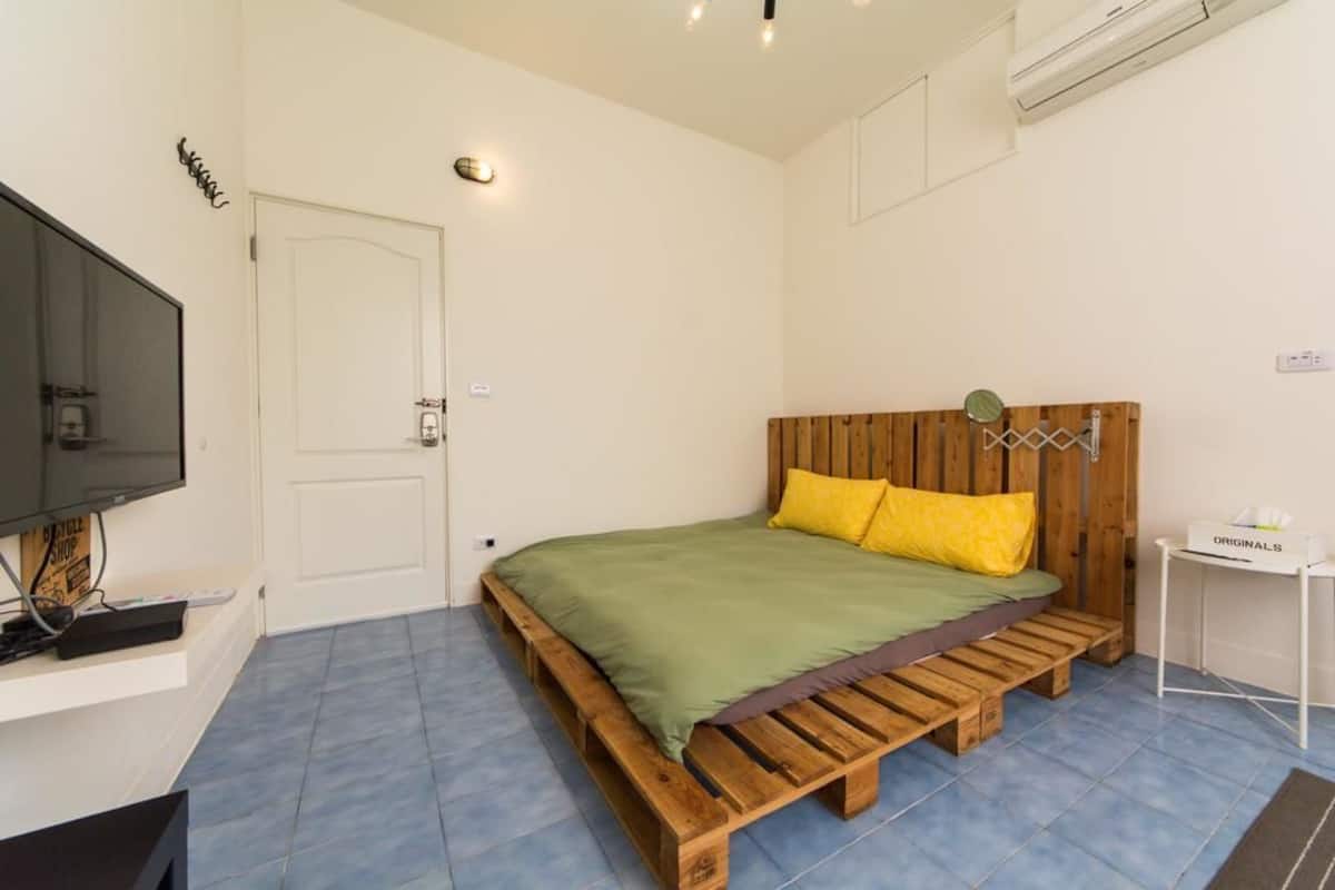 A bedroom with a wooden bed, green bedding, and a small white table with a box labeled "ORIGINALS".
