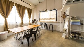 A dining area with a long wooden table, black chairs, and a white chair. There are hanging lights, a window with curtains, and a shelf with folded blankets. - Memory Traveler (Tainan)