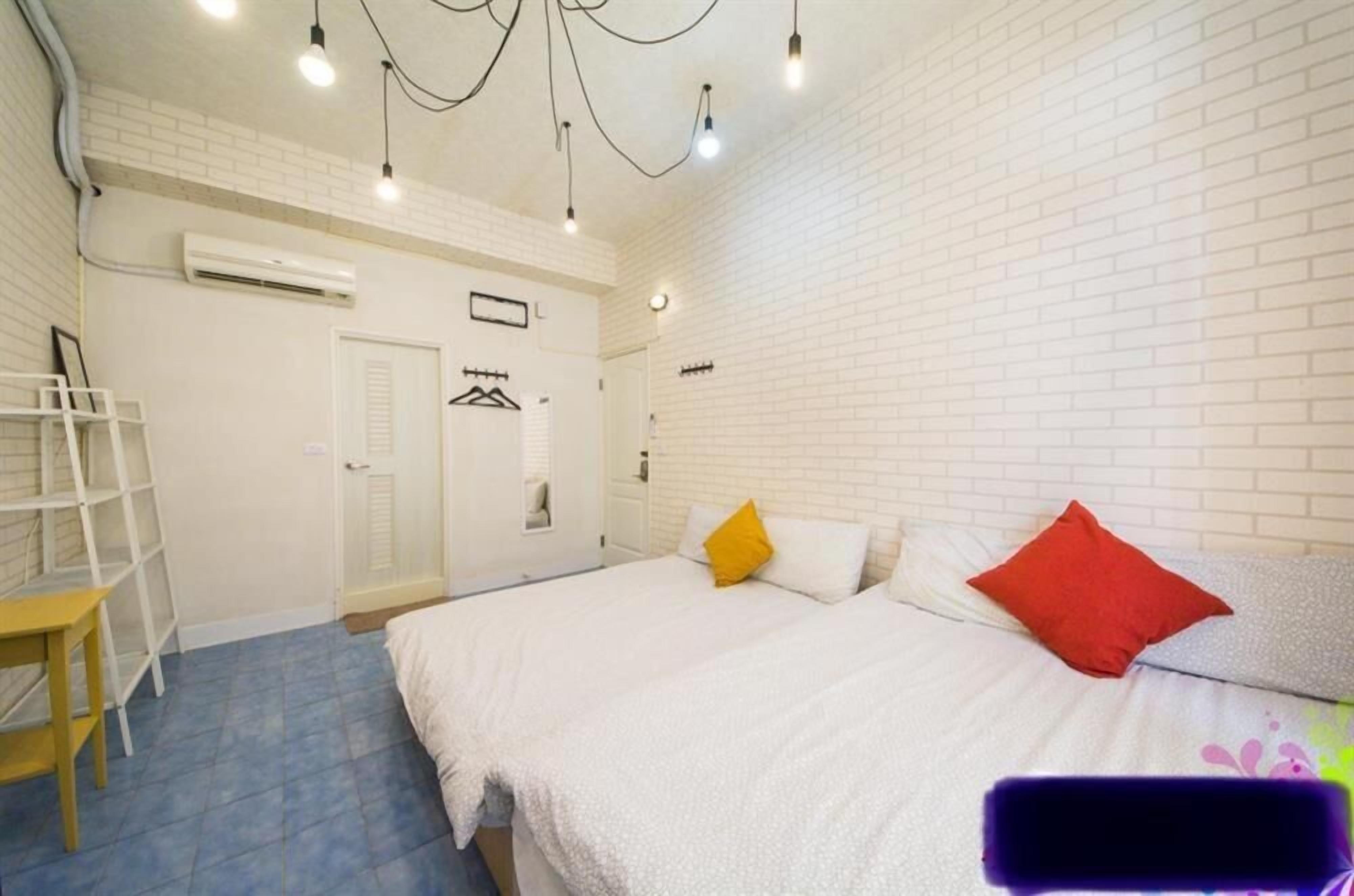 Un dormitorio con pared de ladrillo blanco, cama con ropa blanca y dos almohadas, una mesita pequeña y una lámpara colgante.