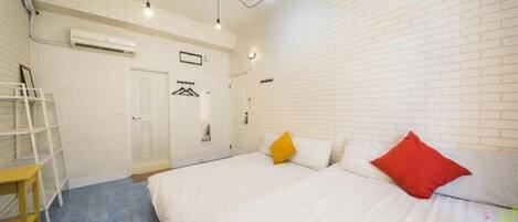 Un dormitorio con pared de ladrillo blanco, cama con ropa blanca y dos almohadas, una mesita pequeña y una lámpara colgante.