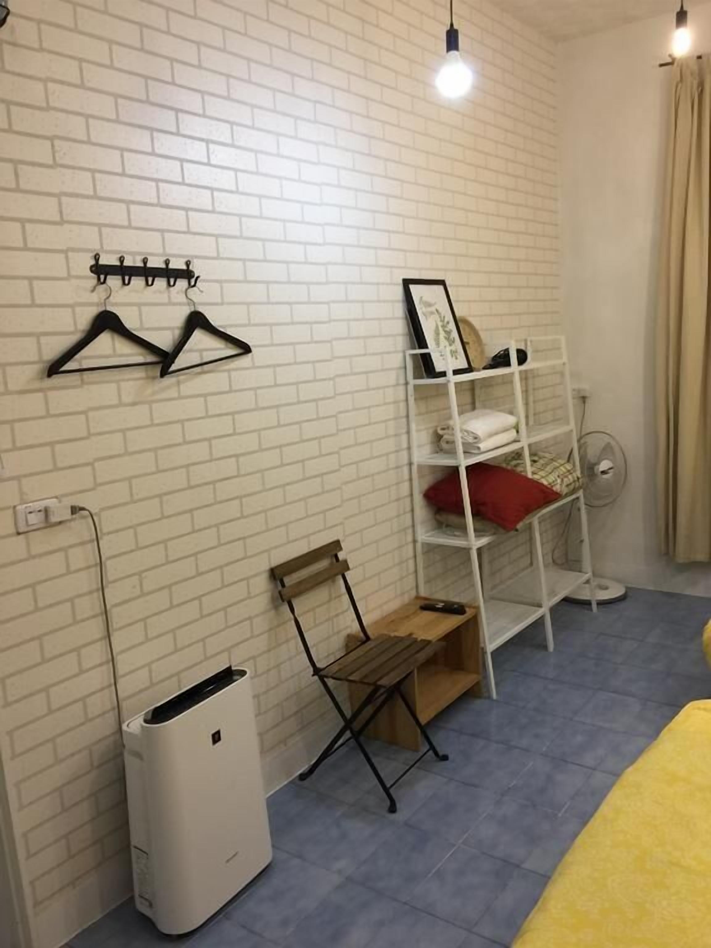 Ett rum med en vit tegelvägg, ett vitt element, en vit hylla med handdukar, en vit papperskorg, en träbänk och en säng med ett gult överkast.