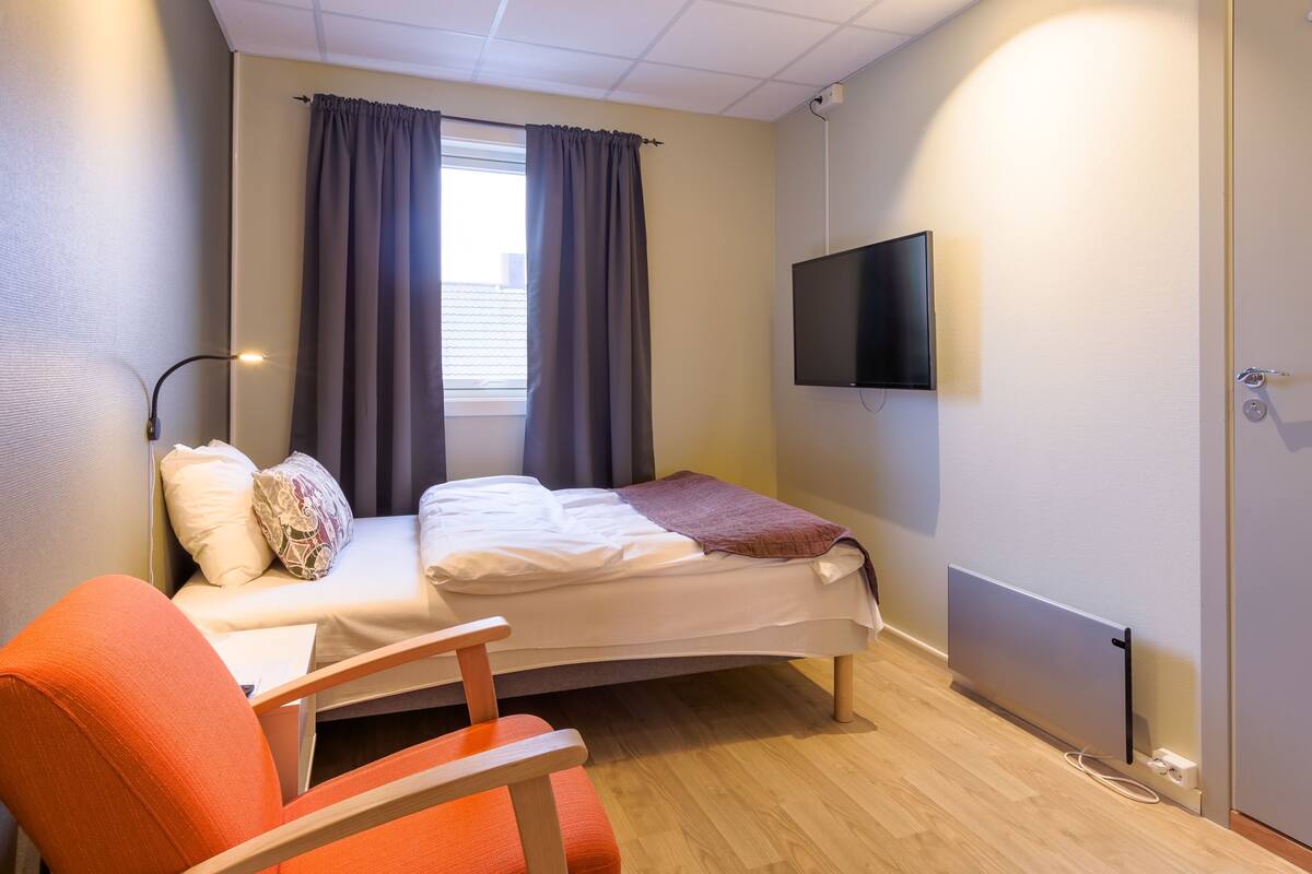 A small, well-lit room with a bed, a chair, a TV, and a window with curtains.