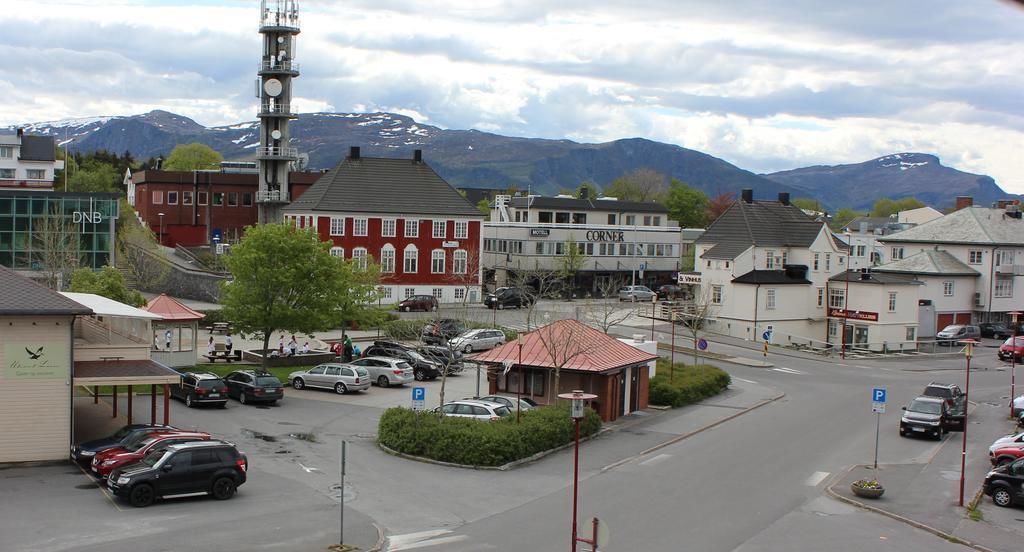 Une place de ville bordée de bâtiments, avec un espace de stationnement et une chaîne de montagnes en arrière-plan.