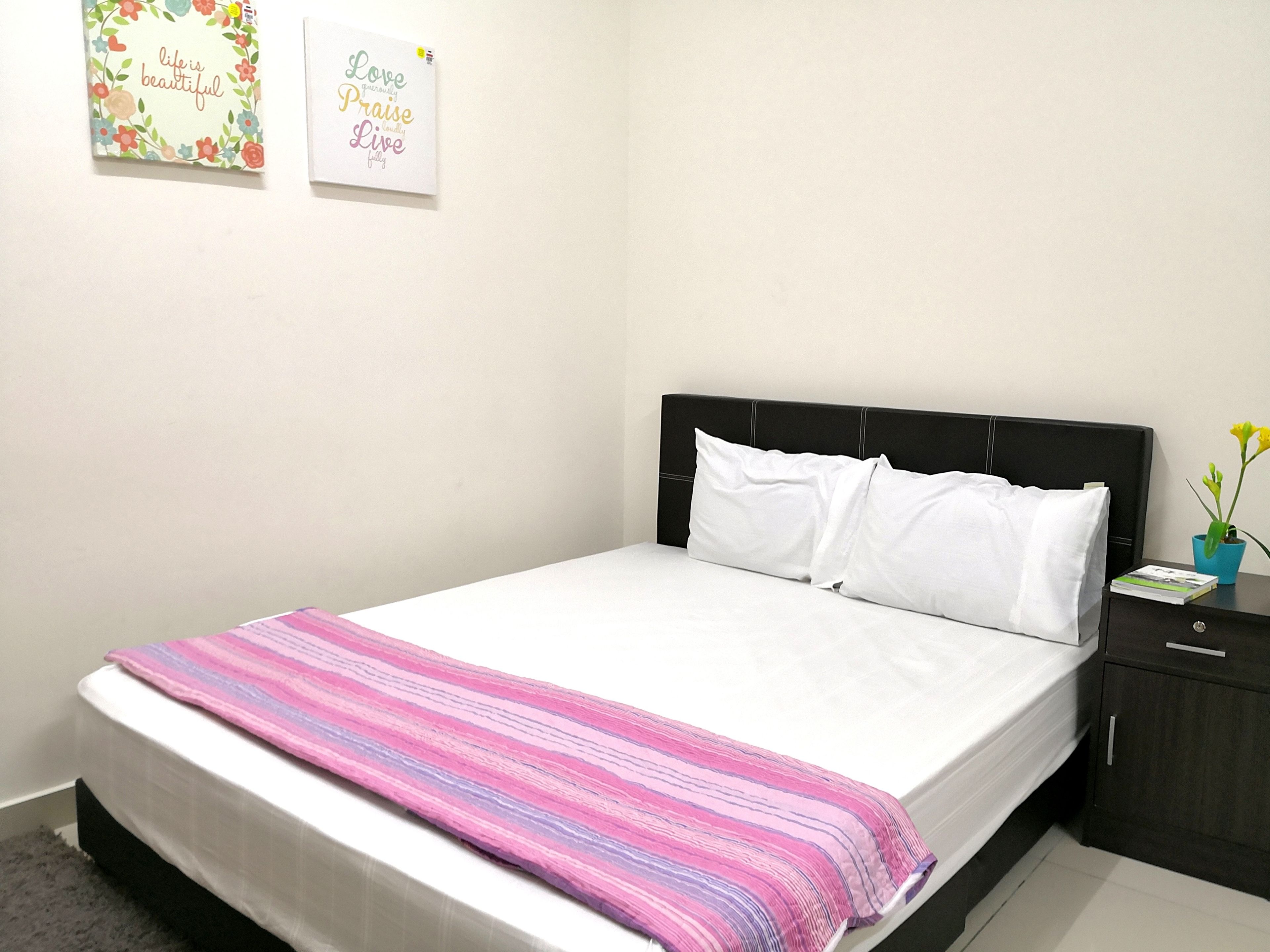 A neatly made bed with white bedding and a pink and purple striped blanket, a black headboard, and a small bedside table with a book and a potted plant.