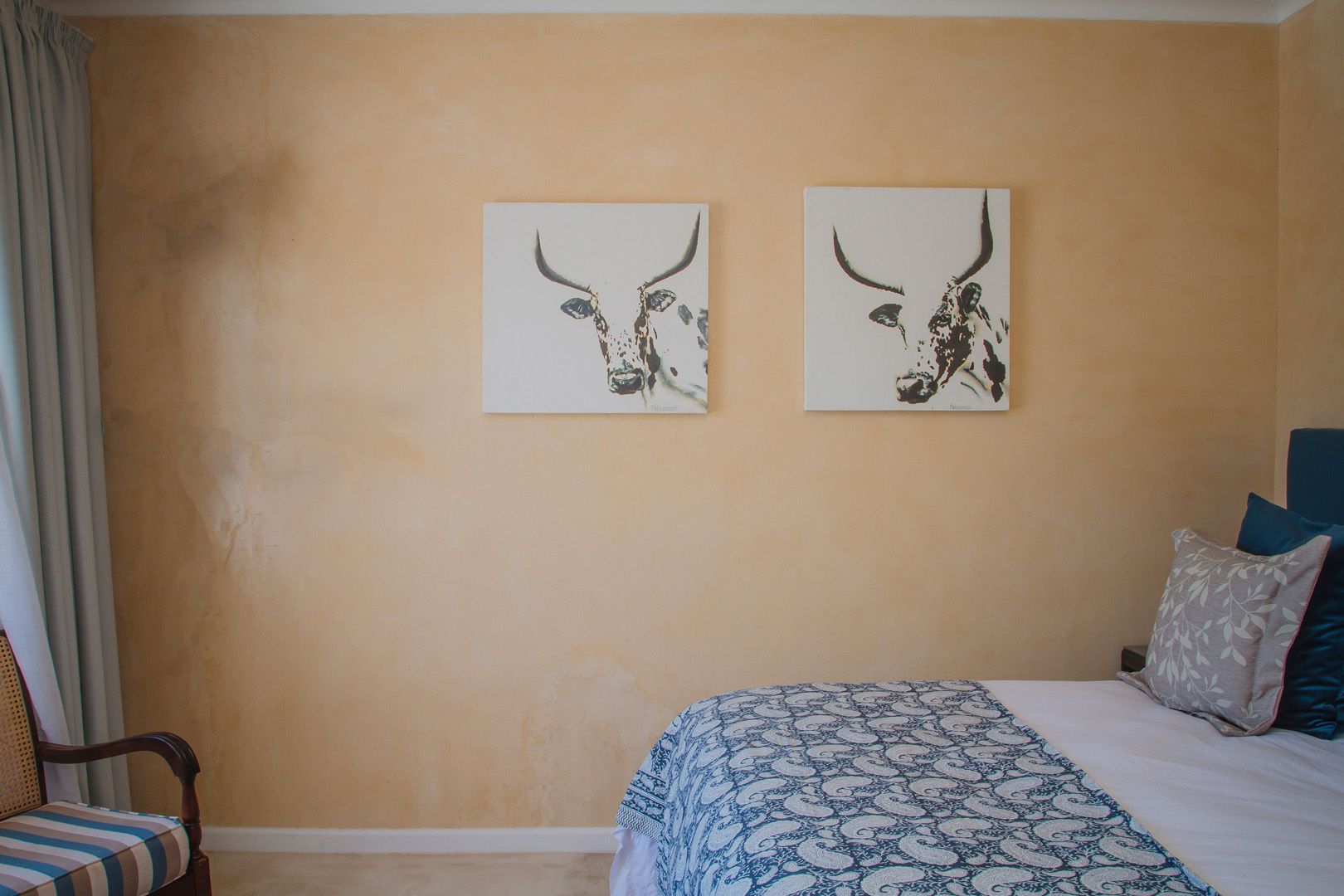 Una camera da letto con una parete color beige, due opere d'arte incorniciate raffiguranti teste di toro, un letto con copripiumino decorato e una sedia con cuscino a righe.