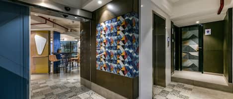 A modern hotel lobby with patterned flooring, a colorful geometric wall, and a seating area with chairs and a table.