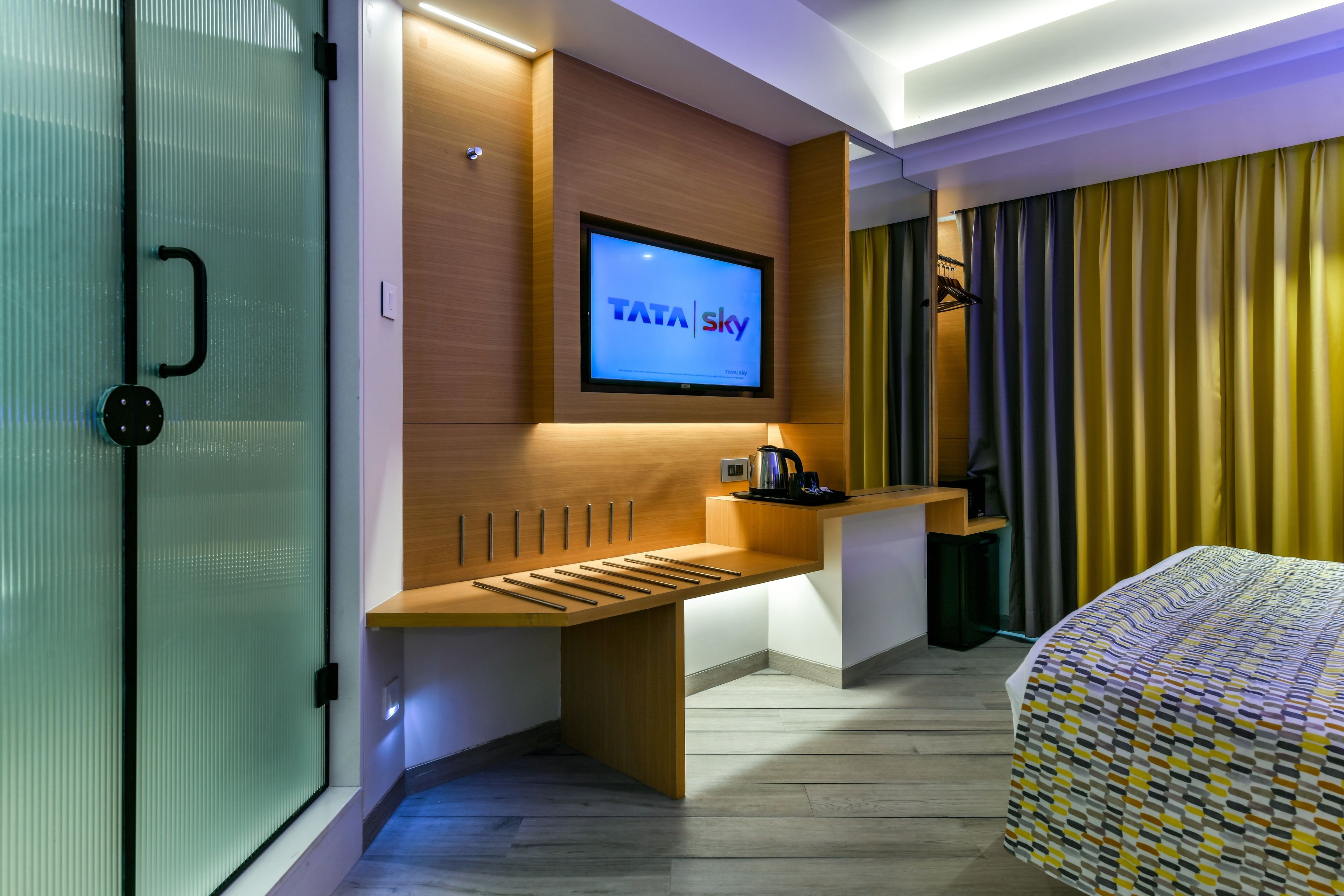 Et moderne hotelværelse med et fladskærms-tv, der viser TATA Sky-logoet, et træskrivebord, en seng med et mønstret sengetæppe og et vindue med gardiner.