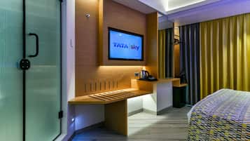 Una habitación de hotel moderna con un televisor de pantalla plana mostrando el logotipo de TATA Sky, un escritorio de madera, una cama con una colcha estampada y una ventana cubierta por cortinas.