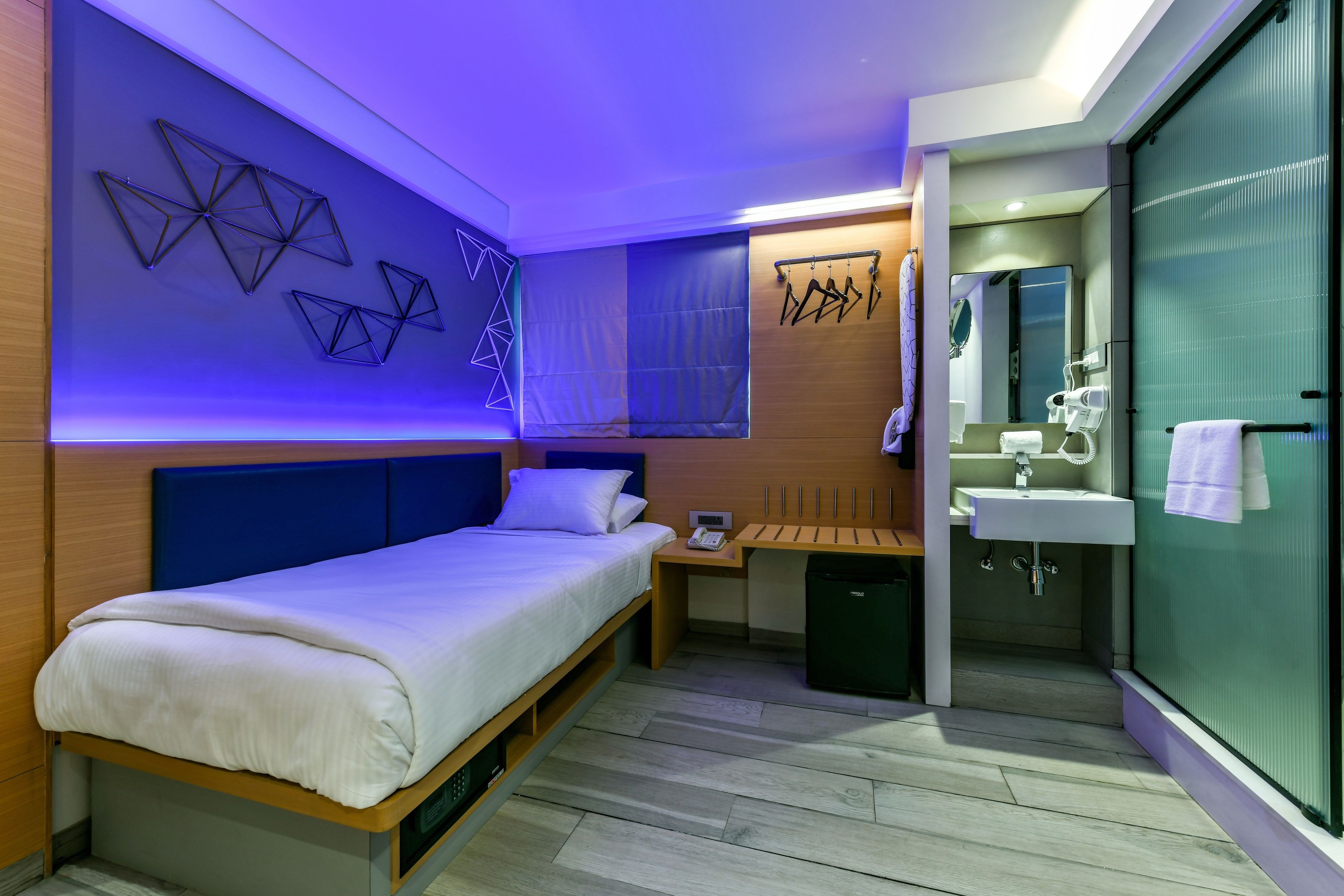 A modern hotel room with a bed, bedside table, and a small desk with a chair. There is a bathroom with a sink and mirror.