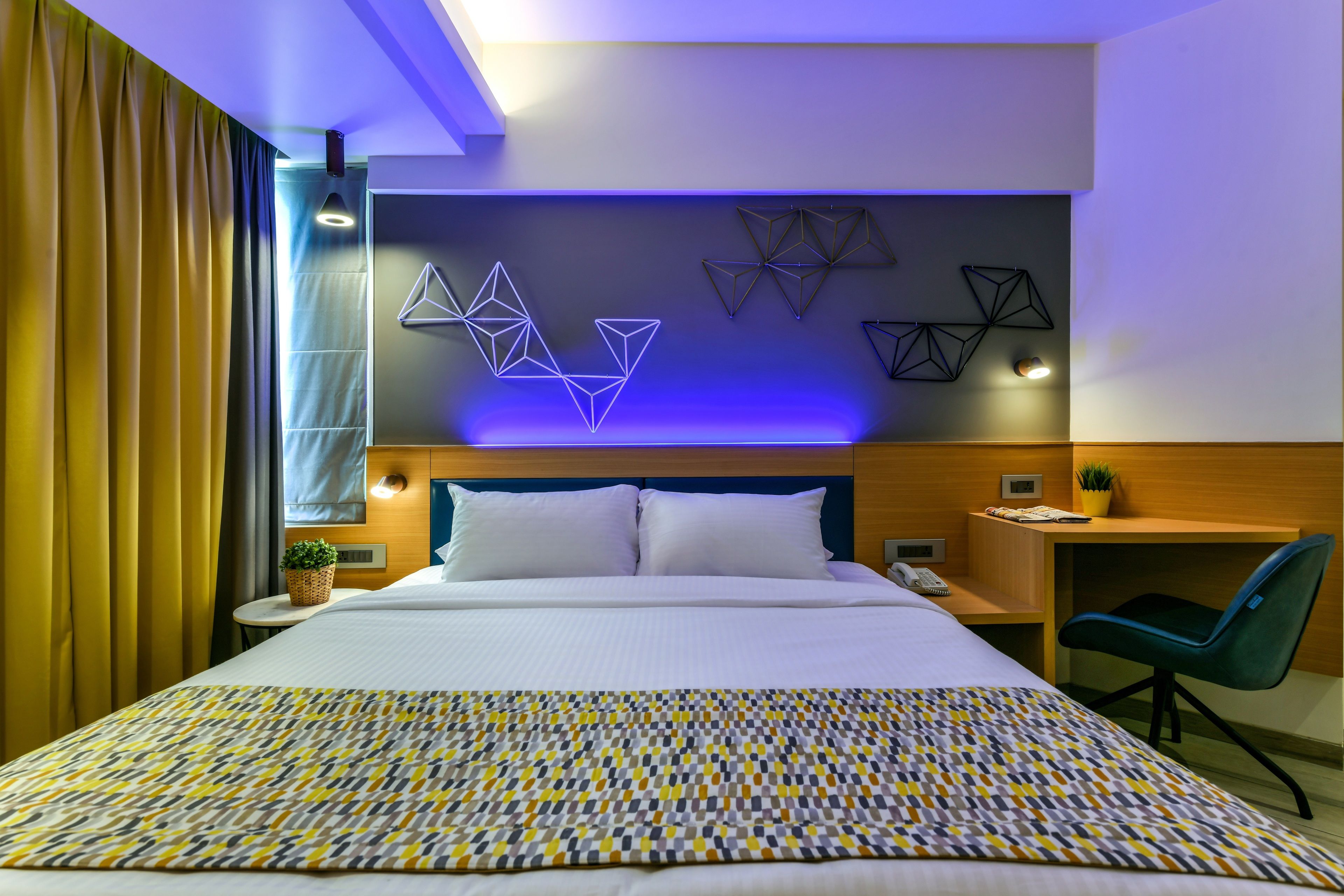 A modern hotel room with a large bed, a desk, a chair, and geometric wall art.