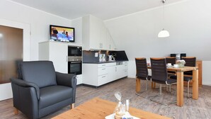 Una sala moderna con un sofá, una mesa de comedor de madera y una cocina con armarios blancos y un televisor montado en la pared.