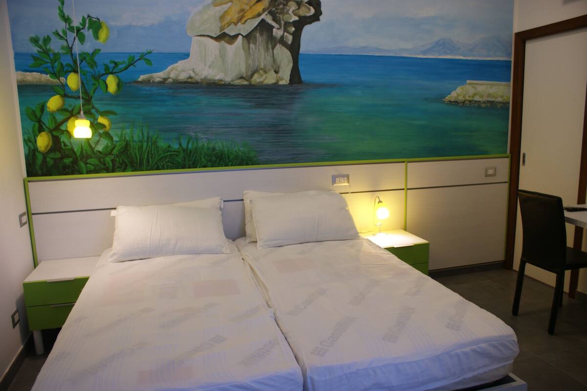 Ein Doppelbett mit weißen Leintüchern, zwei Nachttische, ein Schreibtisch mit Stuhl und ein Wandgemälde mit einer Küstenlandschaft.