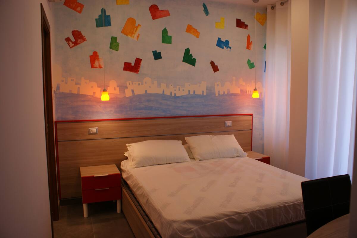 Ein Hotelzimmer mit einem Holz-Kopfteil, einem Bett mit weißen Leinentüchern, einem kleinen Nachttisch und einer Wand mit farbenfrohen ausgeschnittenen Formen.