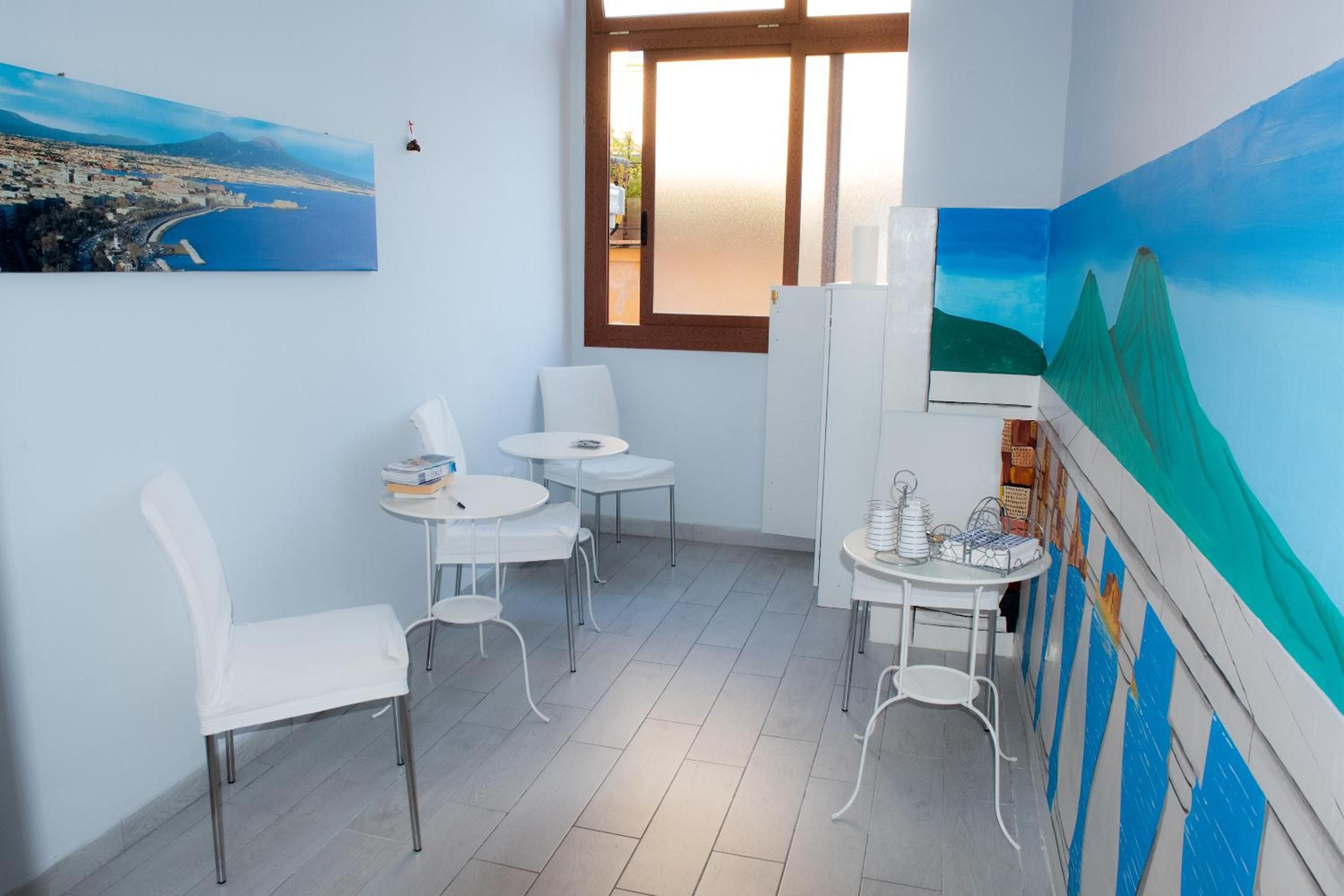 A room with a large window, white chairs, a small table with books, and a painting of a landscape.