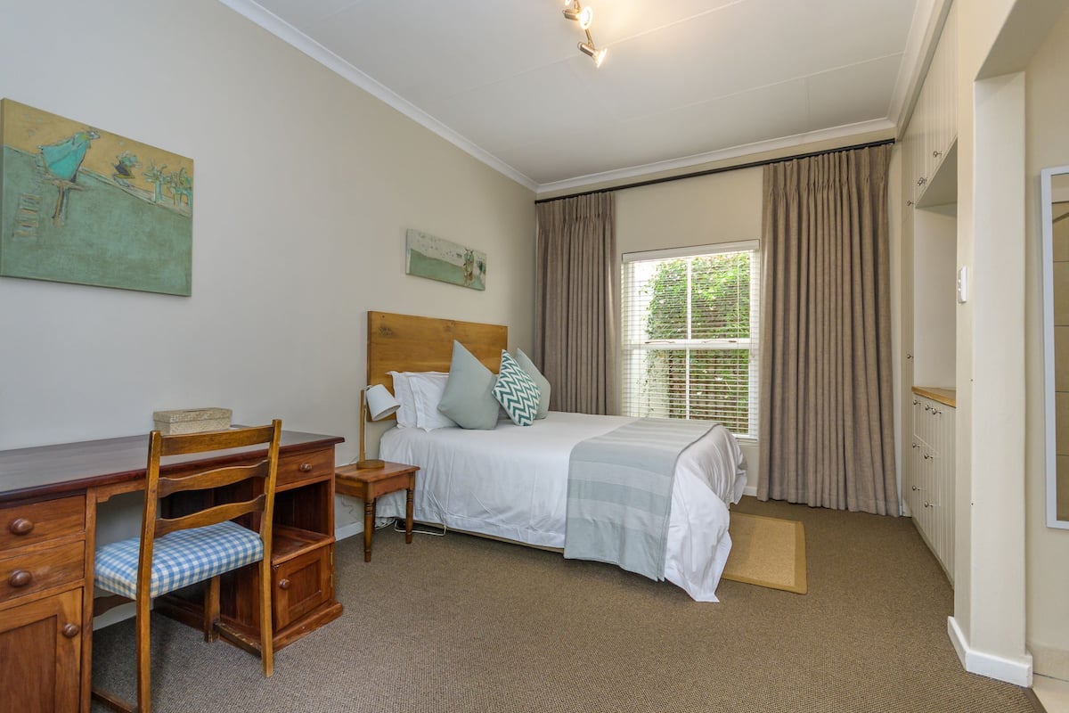 A bedroom with a bed, a desk, and a chair. There is a window with curtains, a painting on the wall, and a door.