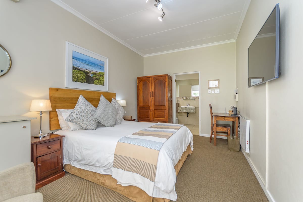 A bedroom with a bed, bedside table, lamp, wooden wardrobe, desk, chair, and a wall-mounted TV.