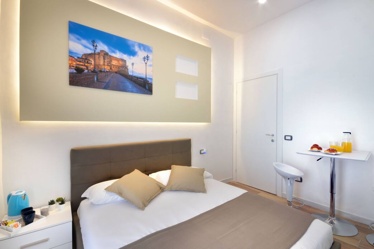 A modern hotel room with a large bed, a bedside table with a coffee maker, a small sink, and a painting of a cityscape on the wall.