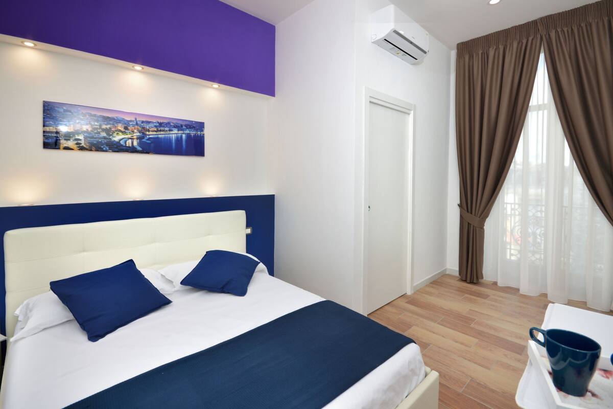 A hotel room with a bed, a painting, a door, and a window with curtains.