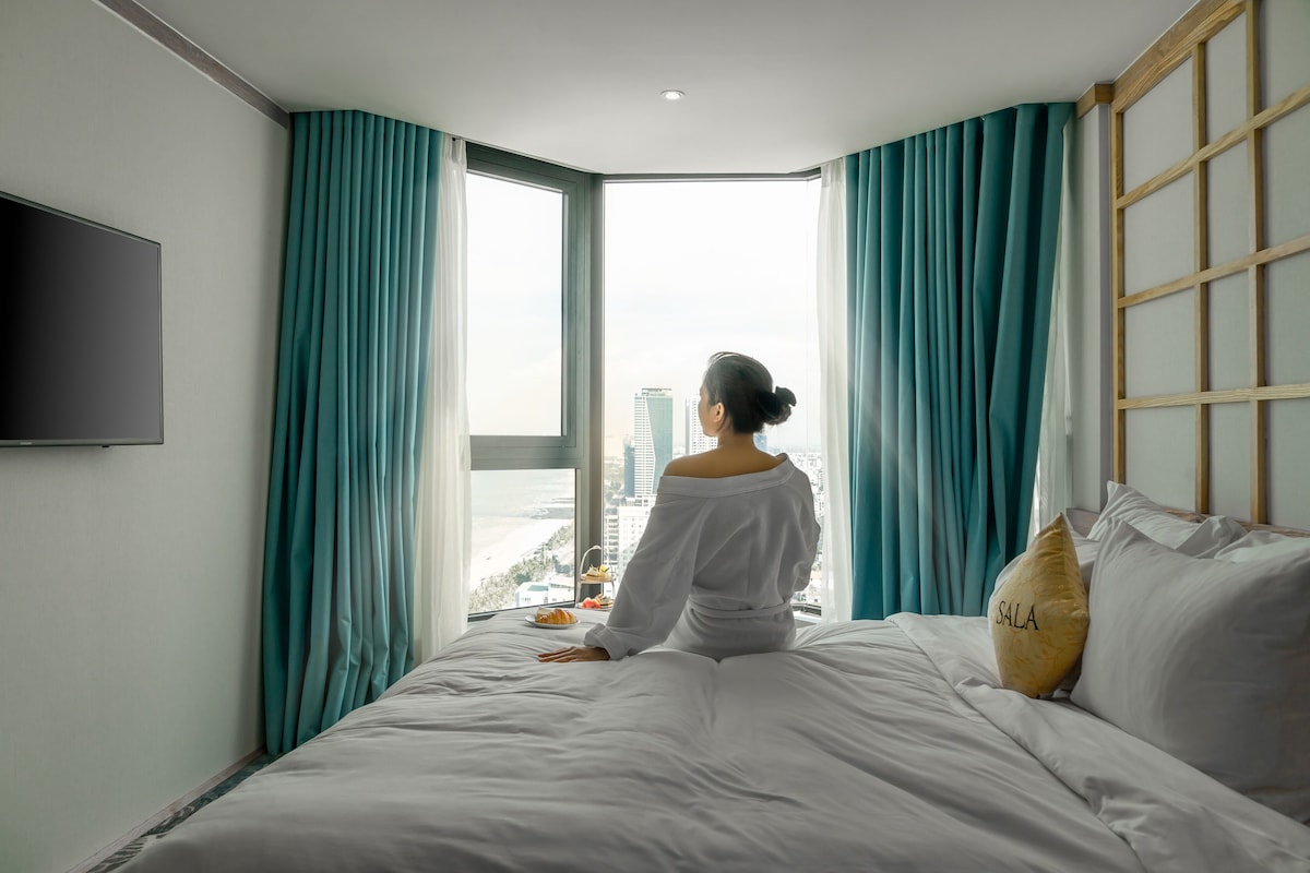 A hotel room with a large window, a bed with white bedding, and a television mounted on the wall.