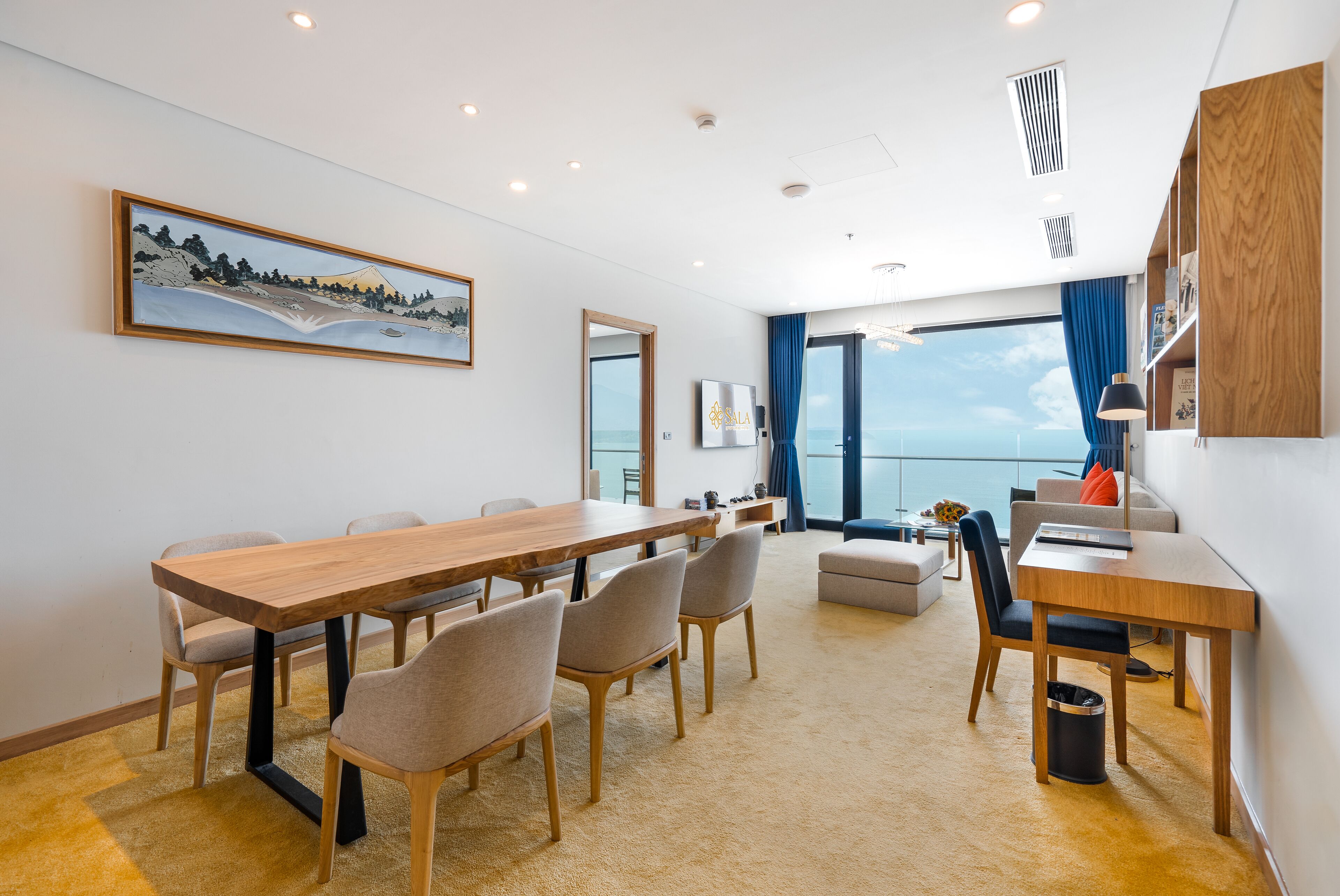 A modern hotel room with a wooden dining table, comfortable seating, a large window with a view, and a mounted artwork.