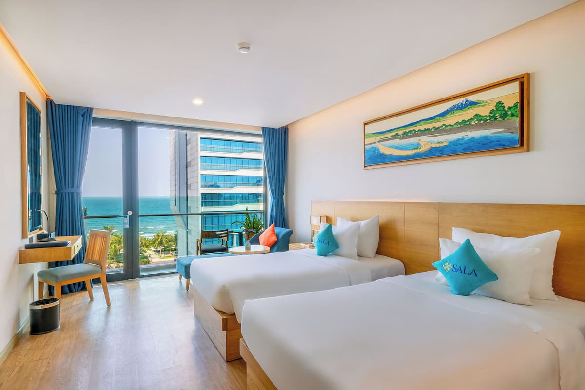 A hotel room with two beds, a desk, a chair, and a view of the ocean.