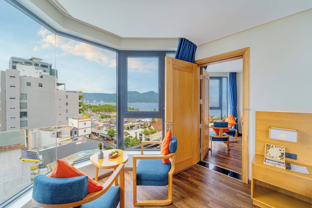 A modern hotel room with a large window offering a city view, a wooden door, and a seating area with blue chairs and a small table.