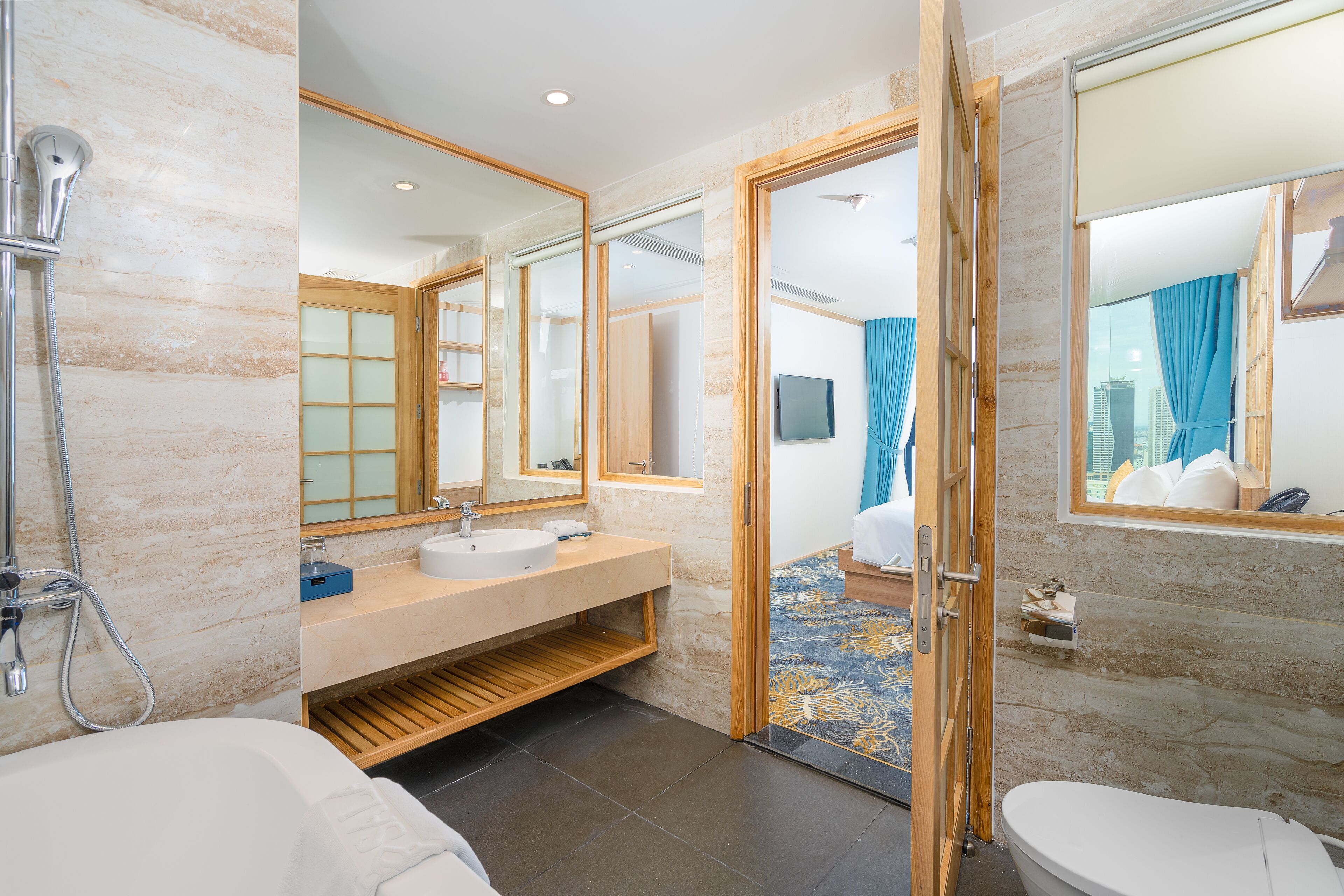 Une salle de bain moderne avec une baignoire, un lavabo et des toilettes. On y trouve une pomme de douche et un miroir au-dessus du lavabo. La salle de bain est dotée d’un plancher en bois et d’un tapis à motifs.
