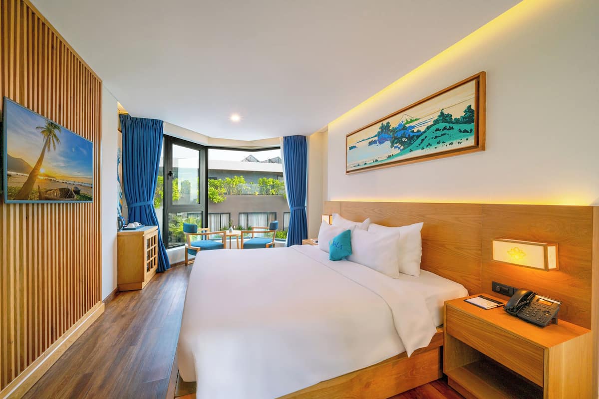 A hotel room with a large bed, a desk with a phone, a painting on the wall, and a view of greenery outside.