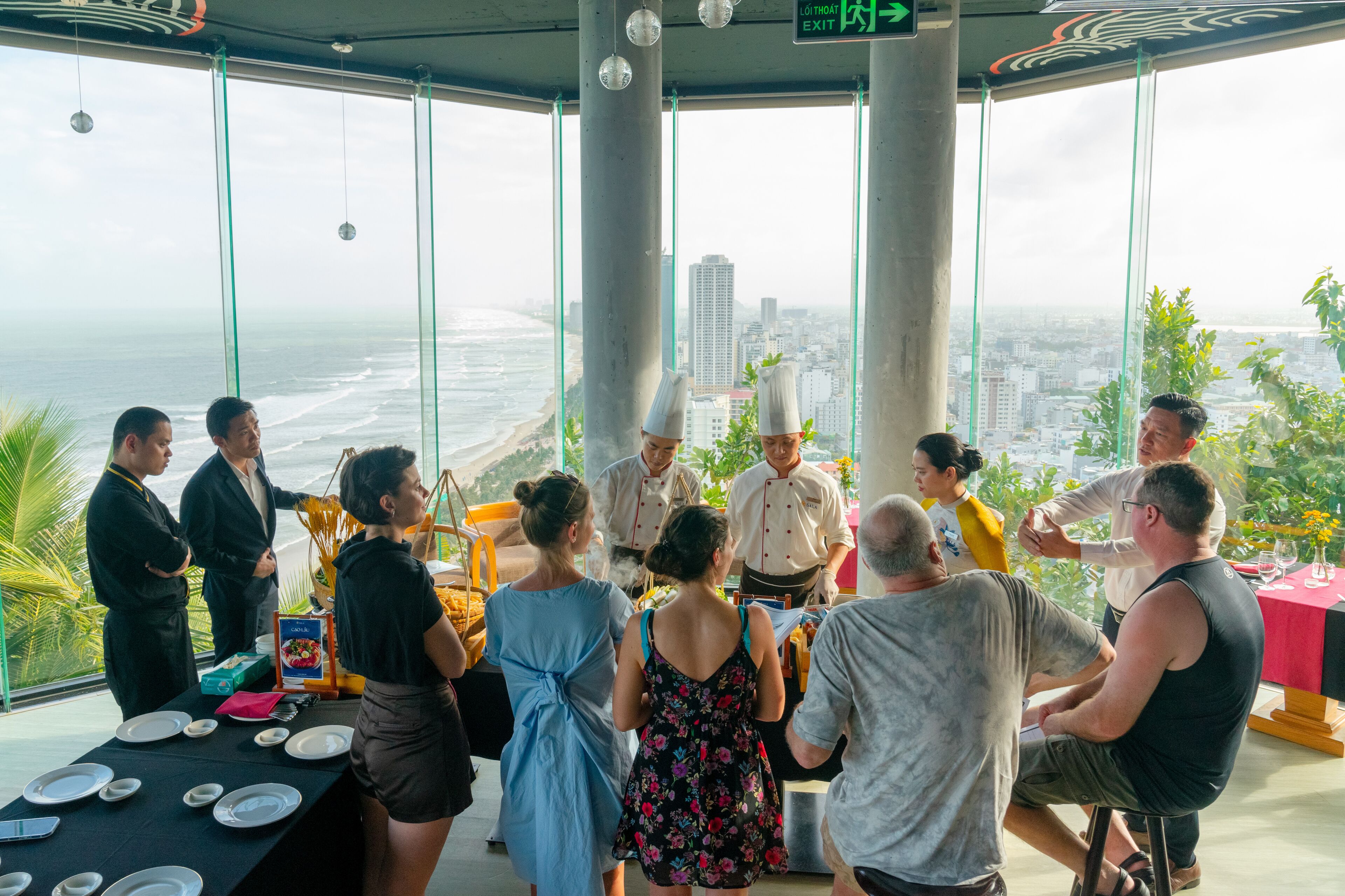 A group of people gathered around a table with a view of the ocean and cityscape through large windows.
